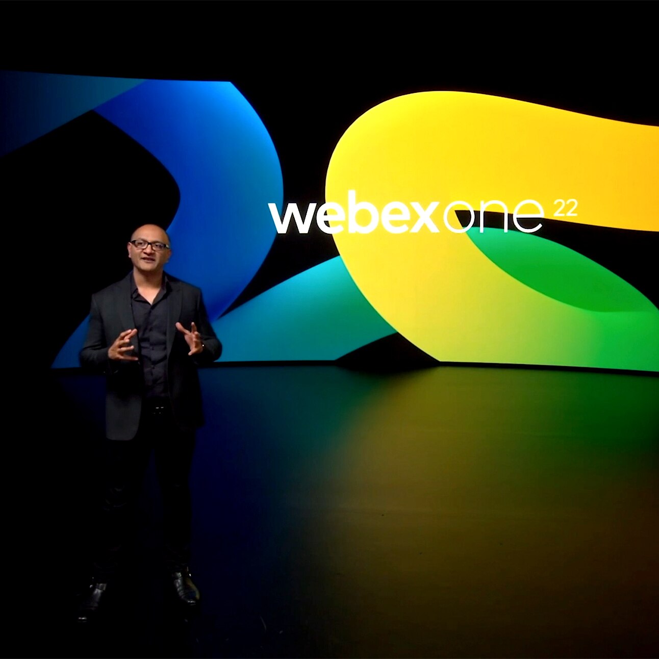 Cisco's Jeetu Patel presents onstage at WebexOne 2022 in front a large black background with a blue, yellow, and green design