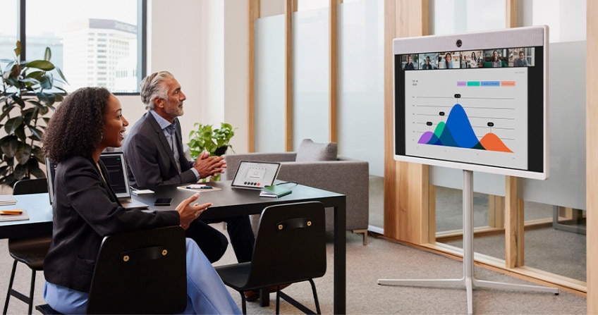 Two colleagues in a conference room video conference with several others. A presentation is displayed on the screen.