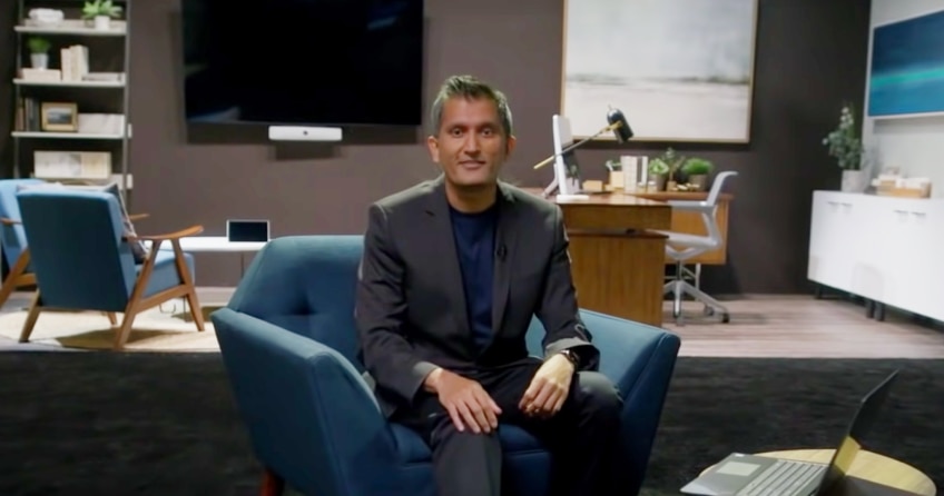 Presenter stands in front of one of the model Webex hybrid workspaces. It contains a desk, armchairs, devices, etc.