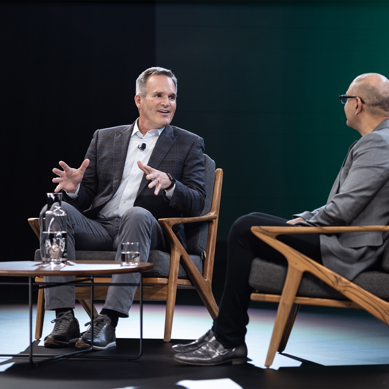 Two speakers chat onstage at WebexOne, seated in wooden chairs around a small, round table.