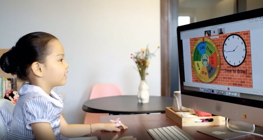 A young child sits at a desk, looking at a computer screen, engaged in virtual learning via video conferencing.
