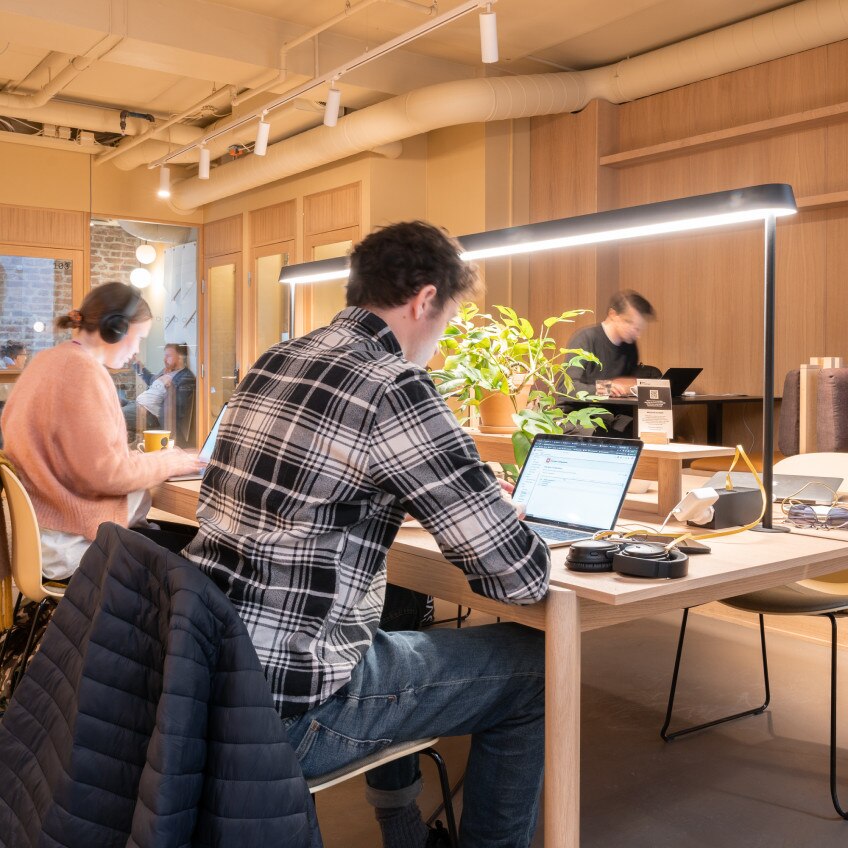 Two people work independently at a large table, in the background others can be seen working at different workspaces.