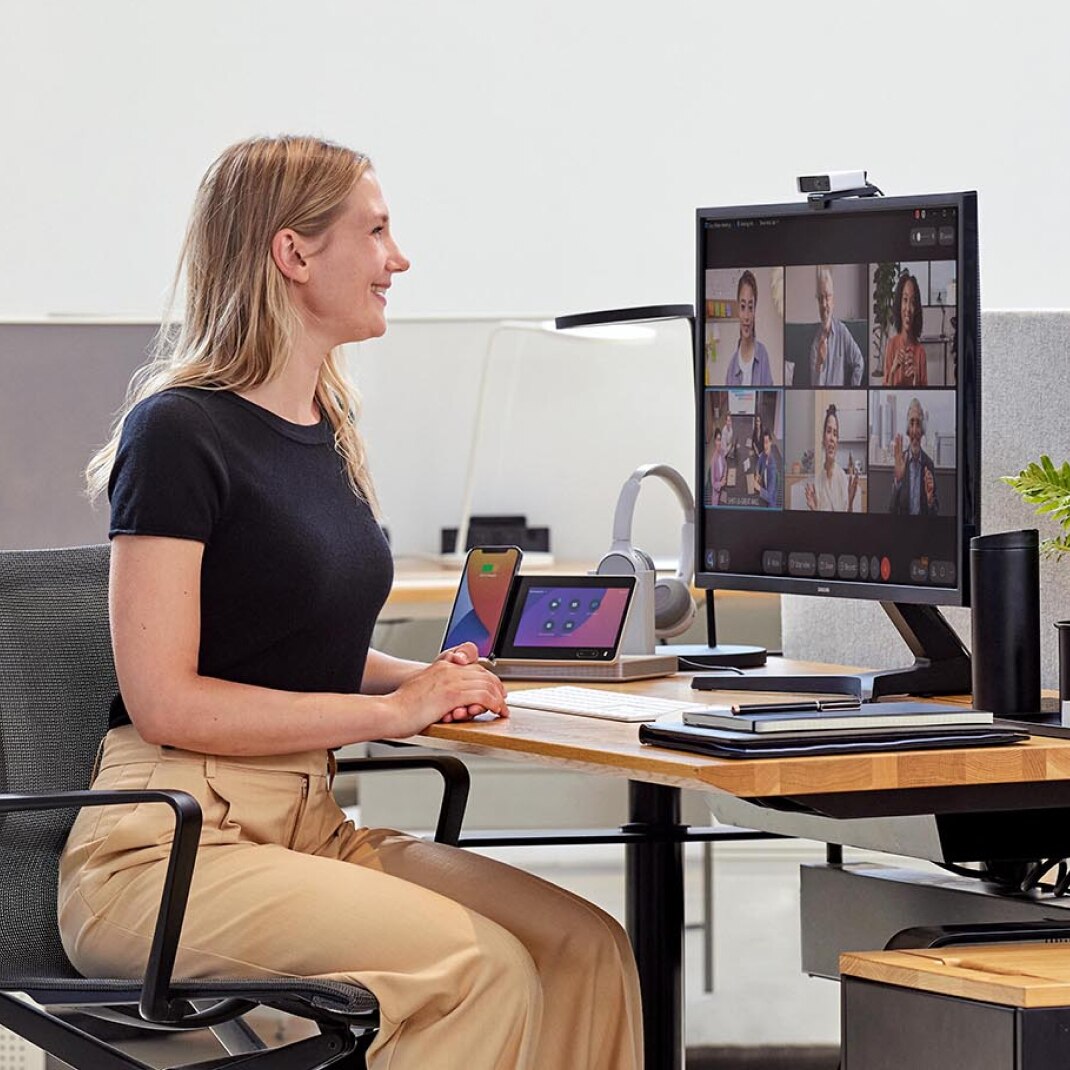 A person wearing a black tee and khaki pants smiles at colleagues in a meeting, which can be seen on a computer monitor.